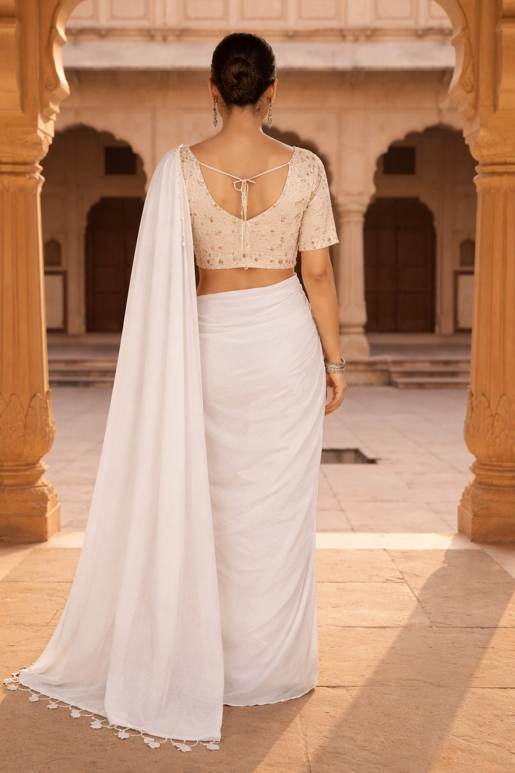 Woman in a traditional outfit with a white saree and beige blouse standing in an architectural setting.